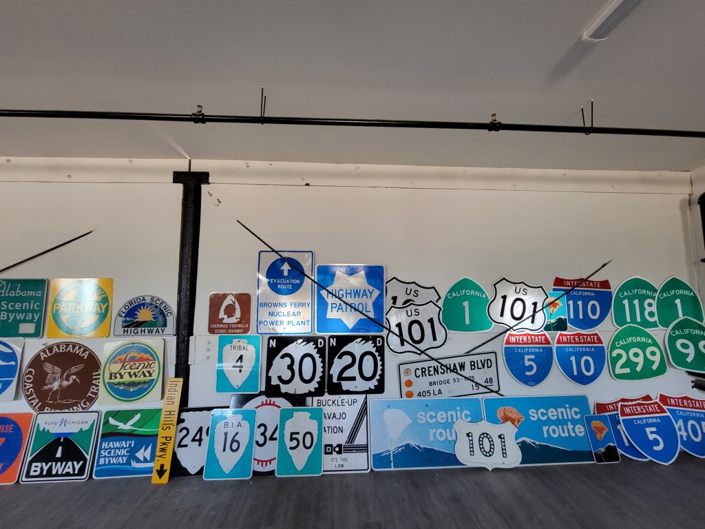 A collection of various road signs displayed on a wall, including highway, scenic route, and state-specific signs, featuring different colors and designs.