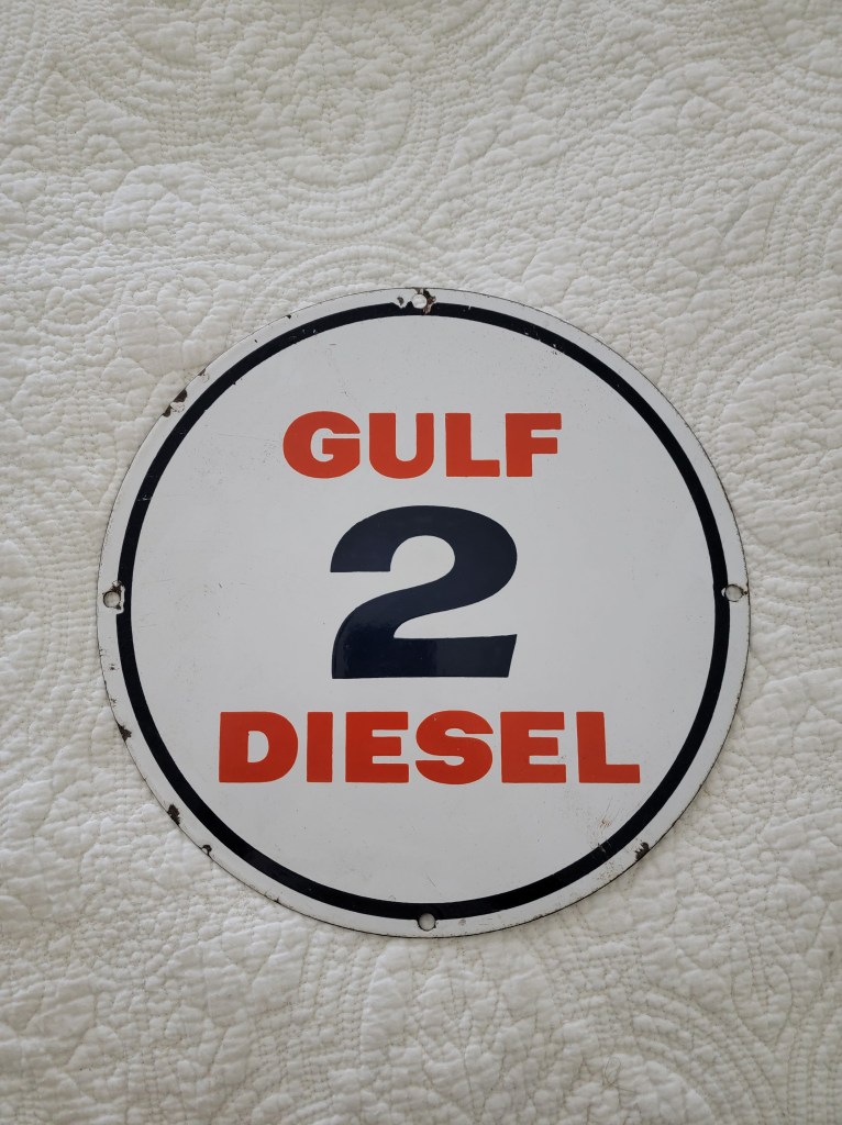 A round Gulf gasoline sign displaying the text '2 DIESEL' in red and black on a white background, resting on a textured white surface. Porcelain