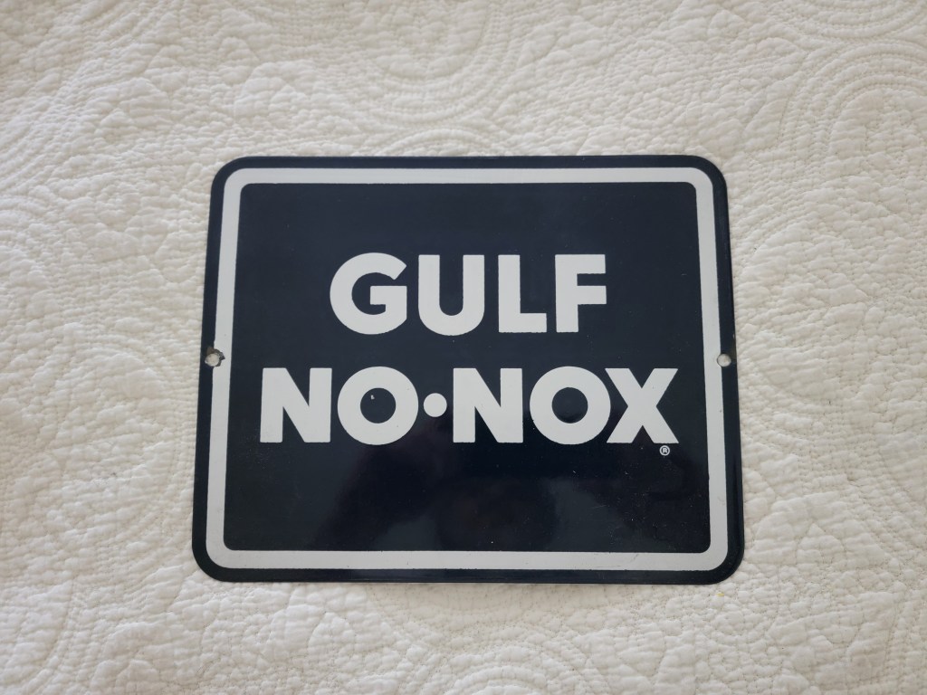 A vintage Gulf No-Nox metal sign featuring bold white letters on a black background, displayed on a textured white quilt.