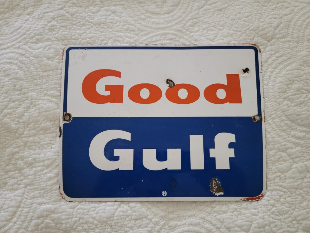A vintage metal sign with the words 'Good Gulf' in orange and white on a blue background, featuring signs of wear and several holes for mounting.