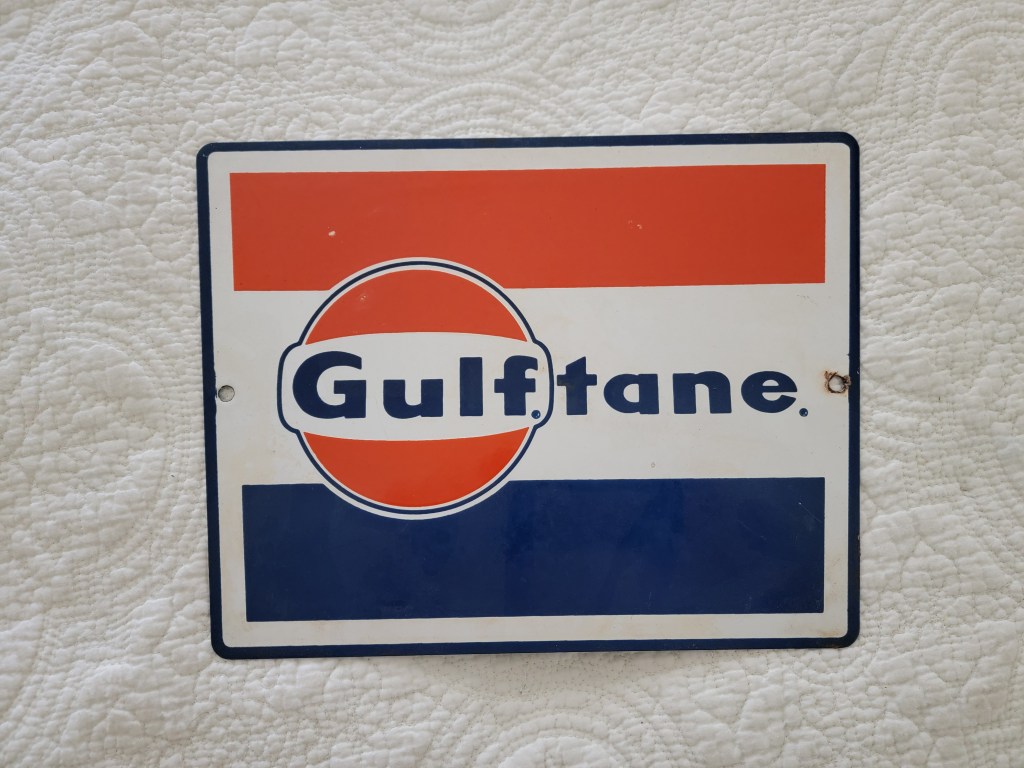 A vintage advertising sign for Gulf oil, featuring the brand name 'Gulf'tane' in bold letters with red, white, and blue colors.