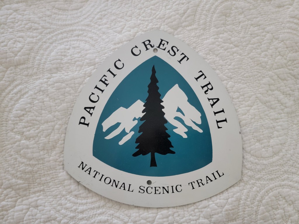 Sign for the Pacific Crest Trail featuring a pine tree and mountains, with the words 'PACIFIC CREST TRAIL' and 'NATIONAL SCENIC TRAIL'.