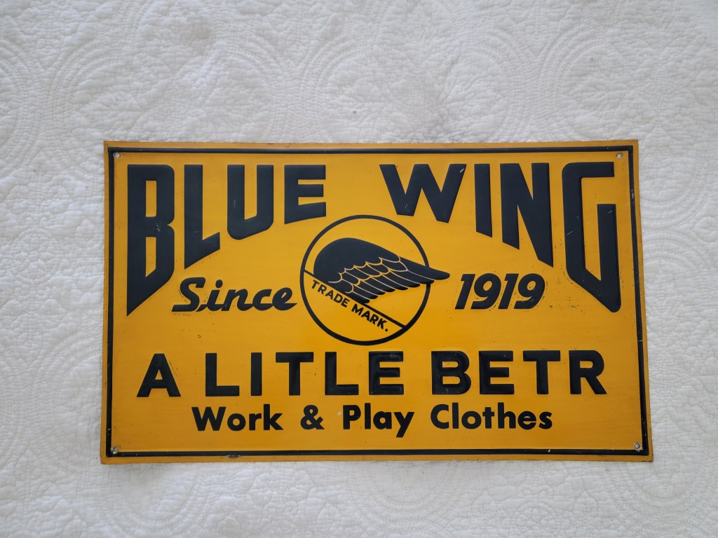 Vintage sign for Blue Wing clothing, featuring the text 'Since 1919' and 'A Little Betr' along with a winged logo on a yellow background.