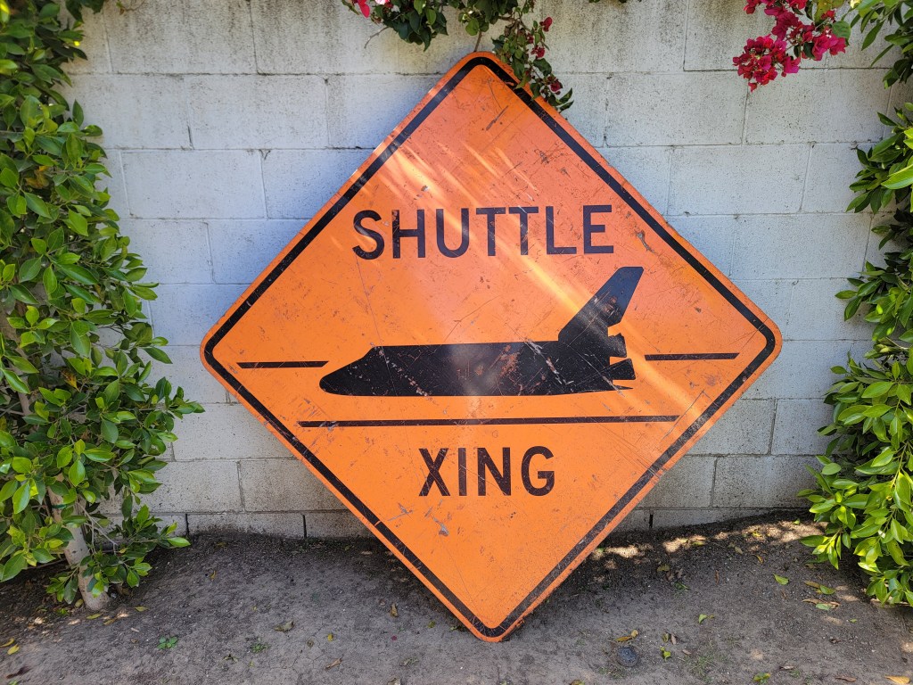 An orange warning sign with the text 'SHUTTLE XING' and an illustration of a space shuttle, surrounded by green foliage and a brick wall.
Rare shuttle crossing sign 4' for the movement of the space shuttle
