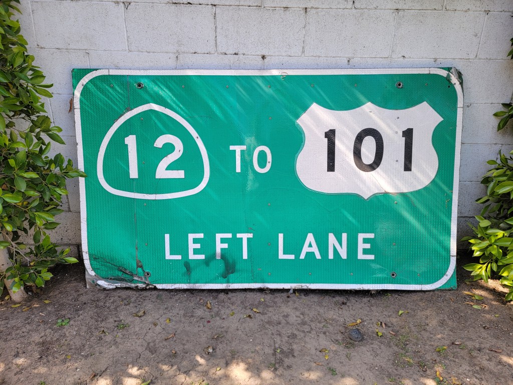 A weathered green highway sign indicating '12 to 101 LEFT LANE' with a shield for route 101, partially obscured by surrounding greenery.
California highway sign 12 to 101 Napa
