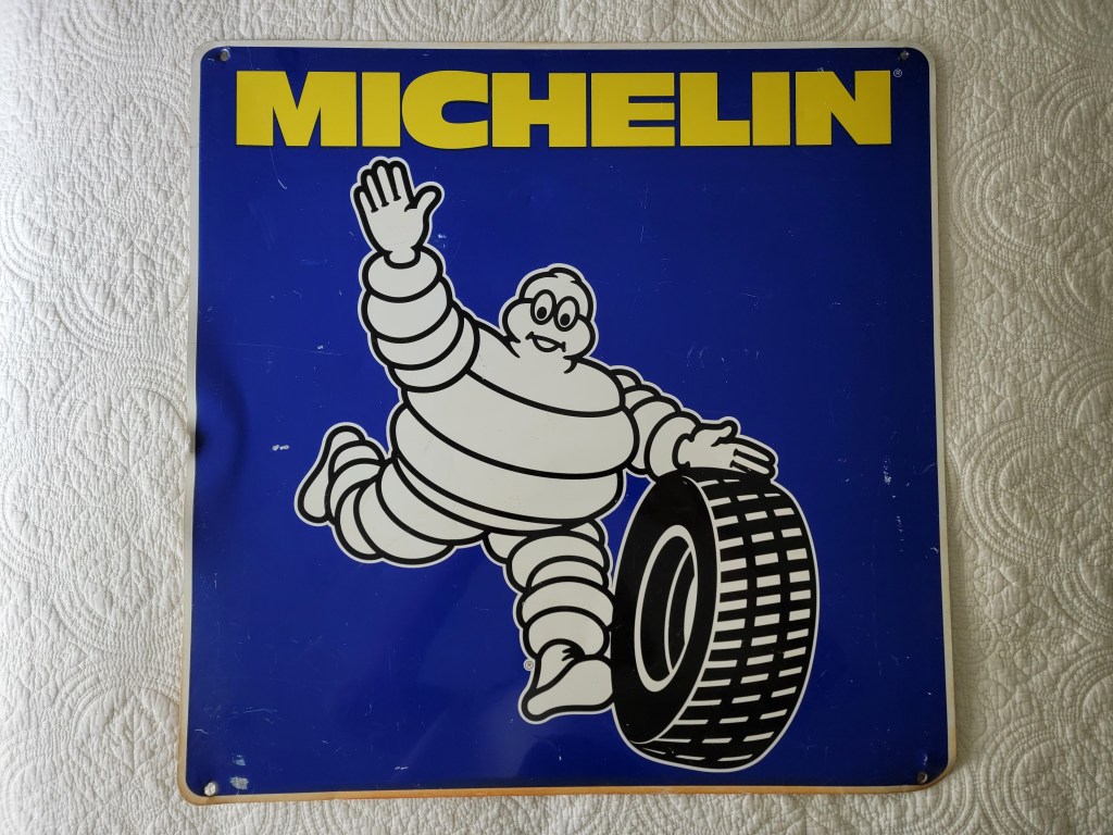 A vintage Michelin sign featuring the iconic Michelin Man character waving while holding a tire against a blue background.