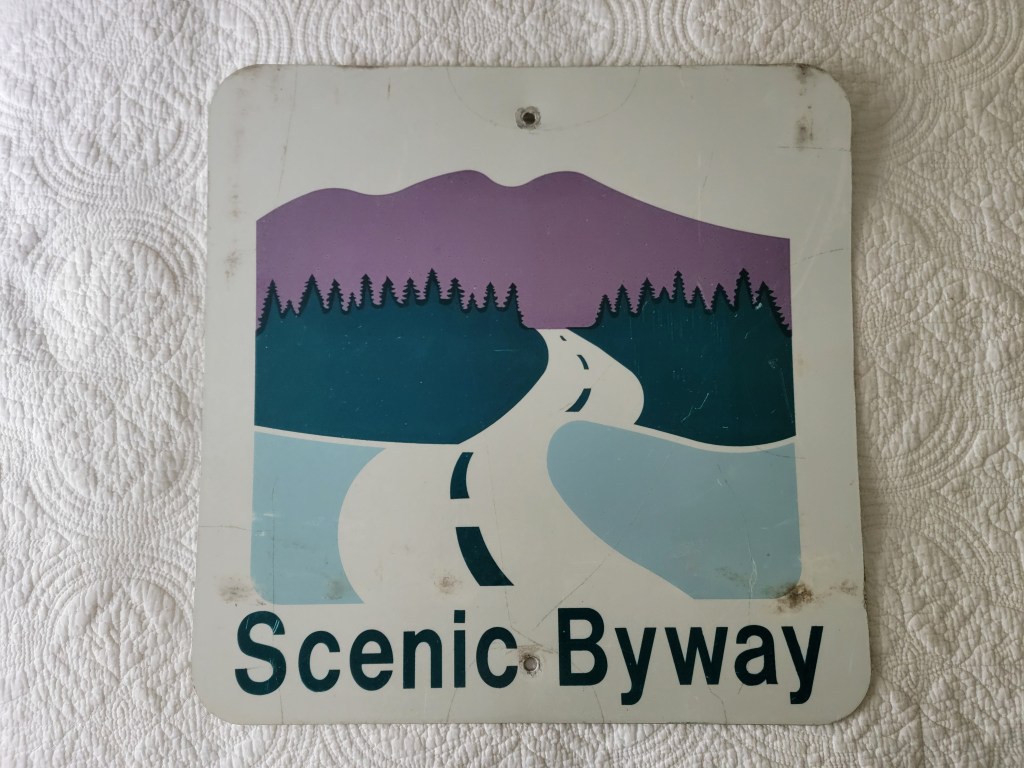 Old Scenic Byway sign featuring a stylized road with mountains and trees in the background, displaying faded colors.
Nation Park Service scenic byway sign
