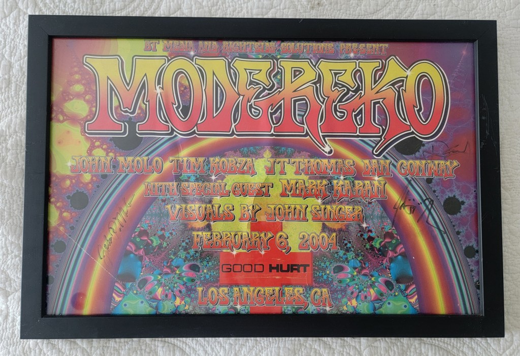 Framed concert poster for Modereko featuring vibrant psychedelic graphics, with names of performers and event details, dated February 6, 2004, in Los Angeles.
Good Hurt Los Angeles California
John Molo, Tim Kobza, JT Thomas, Dan Conway with special Guest Mark Karan Visuals by John Singer
Signed