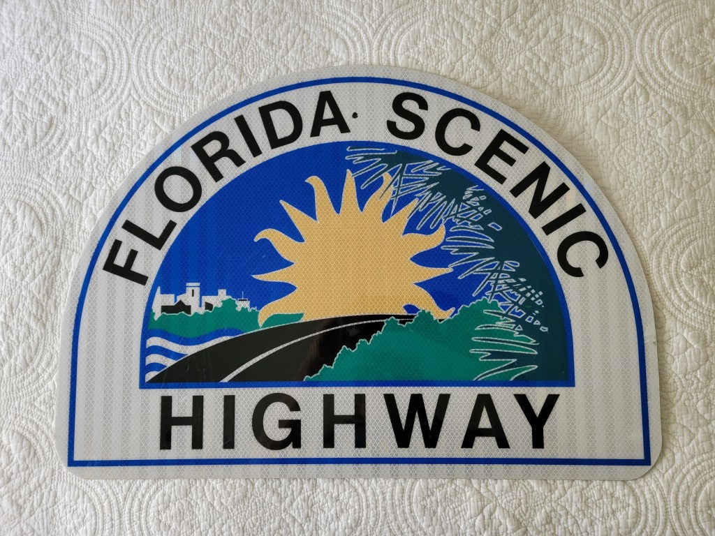 A decorative sign for the Florida Scenic Highway featuring a sun, palm trees, and a road illustration.
