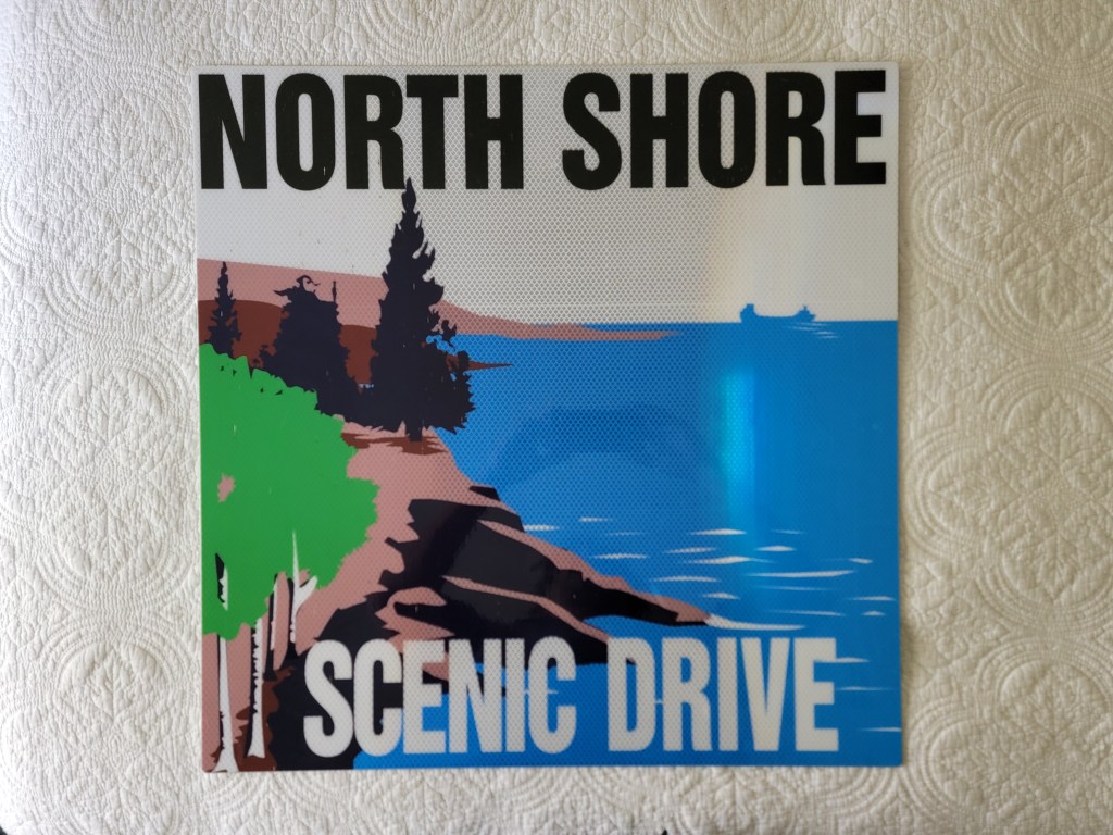 Sign featuring bold text reading 'NORTH SHORE SCENIC DRIVE' with a graphic of trees, rocks, and water in the background.
Minnesota North Shore Scenic Drive Sign