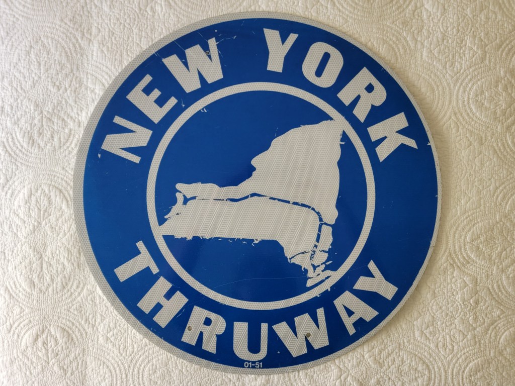 Round blue sign with white text that reads 'NEW YORK THRUWAY' and a silhouette map of New York State in the center.
New York Thruway Sign 1951