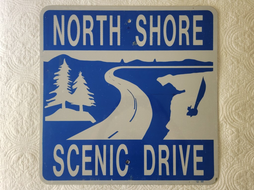 A blue and white road sign that reads 'NORTH SHORE SCENIC DRIVE' with illustrations of trees and a winding road.
Minnesota North Shore Scenic Drive Sign