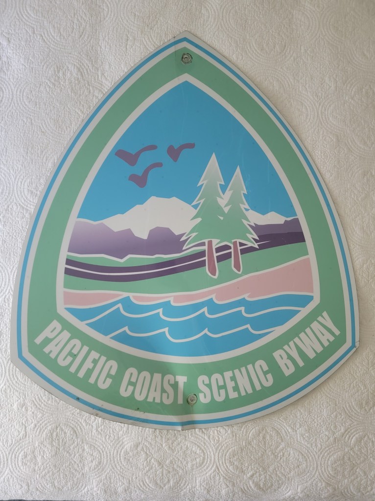 A colorful sign representing the Pacific Coast Scenic Byway, featuring mountains, trees, and water, all in a stylized design.
Washington State Pacific Coast Scenic Byway PCH route 1 sign