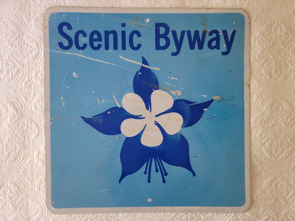 A weathered blue sign reading 'Scenic Byway' with a white flower design.
Colorado Scenic Byway Sign