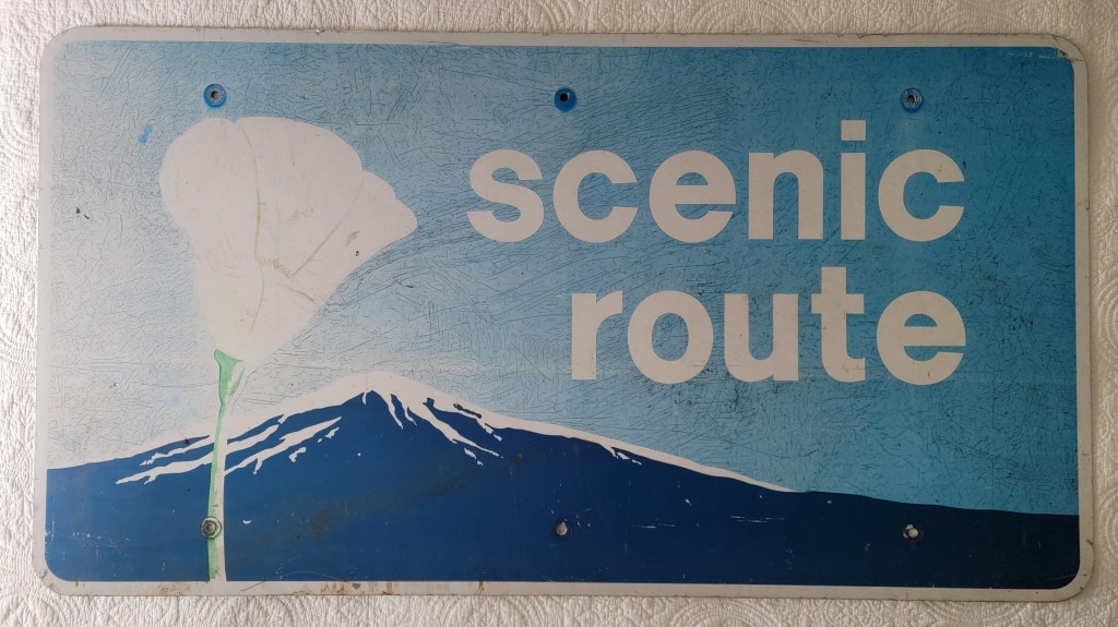 A weathered blue sign with the words 'scenic route' and a white flower illustration alongside a mountain silhouette.
California PCH pacific coast highway poppy sign large street sign