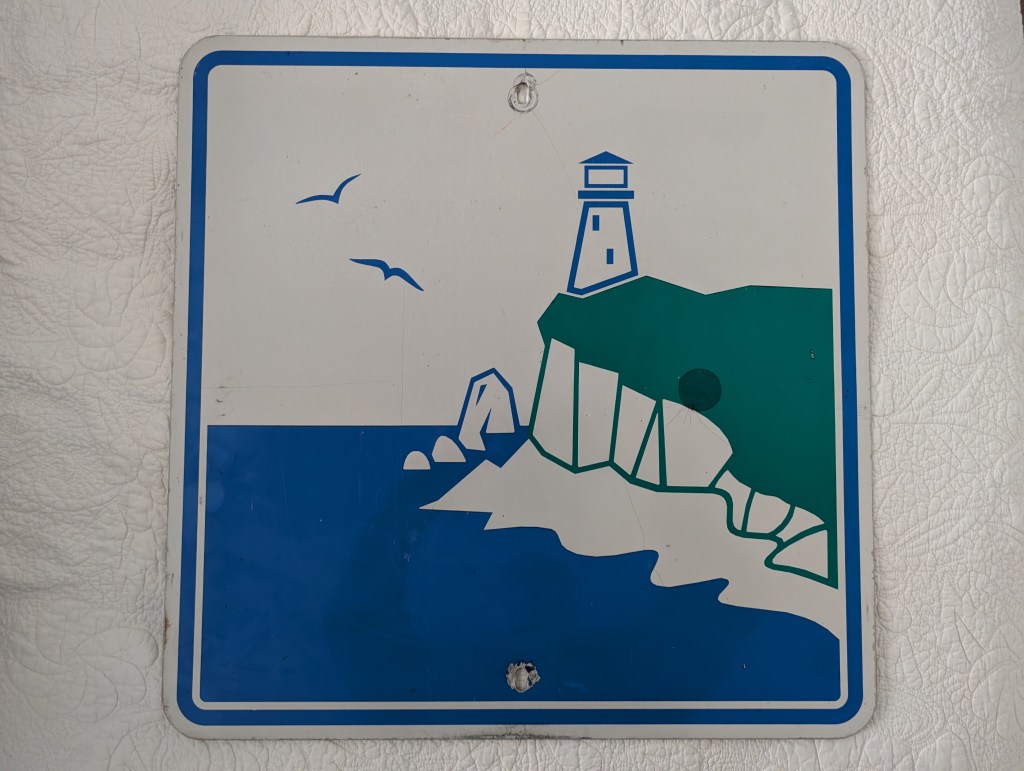 A blue and white sign featuring a lighthouse on a cliff with a rocky shoreline and seagulls flying above.
strret sing